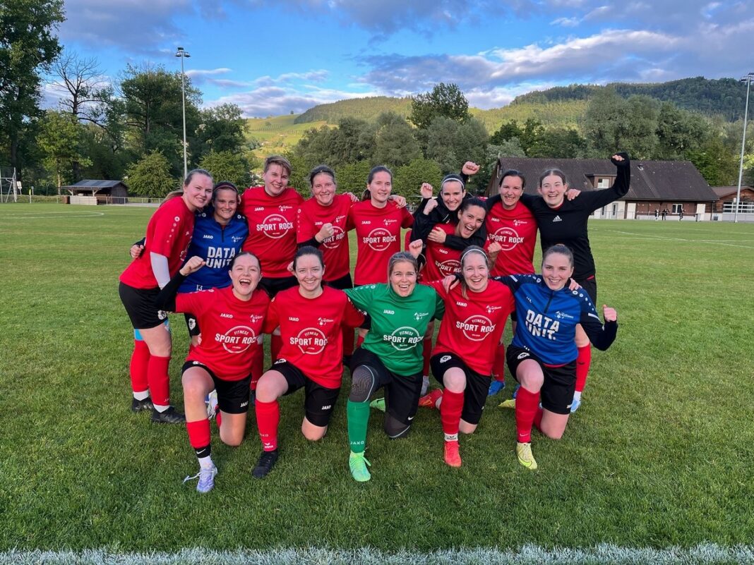 SG-Root-Adligenswil-FC-Willisau Group of women soccer players posing together on a grassy field after a game, wearing red jerseys and cheering with fists raised, hills in the background.