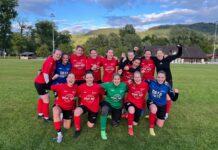 FC Willisau: Geduldsprobe zum Saisonstart Group of women soccer players posing together on a grassy field after a game, wearing red jerseys and cheering with fists raised, hills in the background.