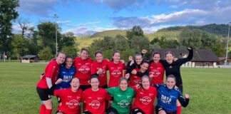Group of women soccer players posing together on a grassy field after a game, wearing red jerseys and cheering with fists raised, hills in the background.