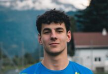 Young man wearing a blue and green sports jersey stands outdoors on a field, mountains in the background, looking at the camera.