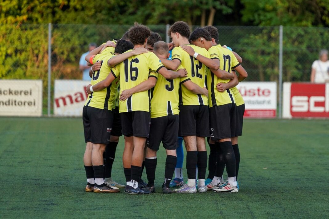 Soccer players in yellow jerseys form a tight huddle on a green turf field, arms around each other before the game.