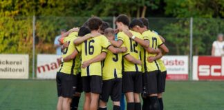 Soccer players in yellow jerseys form a tight huddle on a green turf field, arms around each other before the game.