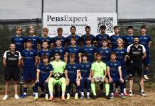 Gelungener Kickoff-Day in Hitzkirch – Seetaler Fussball bereit für die neue Saison Group portrait of a youth soccer team in blue jerseys with two neon-green goalkeepers, two coaches, in front of a PensExpert banner on a chain-link fence.