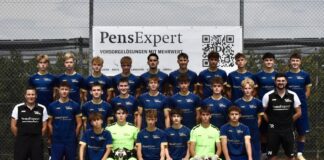 Group portrait of a youth soccer team in blue jerseys with two neon-green goalkeepers, two coaches, in front of a PensExpert banner on a chain-link fence.