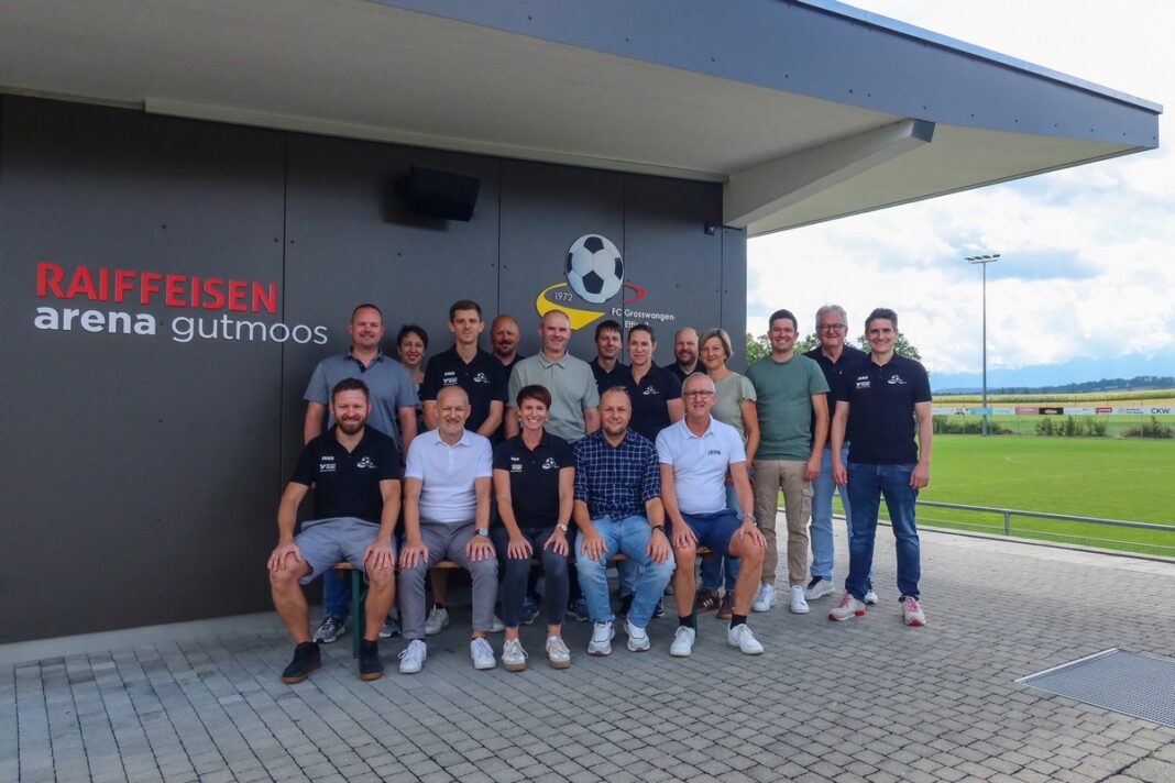 Group of staff posing for a team photo outside Raiffeisen Arena Gutmoos, with a football field in the background.
