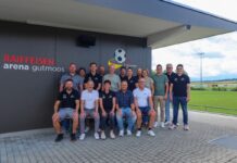 Aus Sportplatz Gutmoos wird Raiffeisen Arena Gutmoos Group of staff posing for a team photo outside Raiffeisen Arena Gutmoos, with a football field in the background.