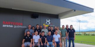 Group of staff posing for a team photo outside Raiffeisen Arena Gutmoos, with a football field in the background.