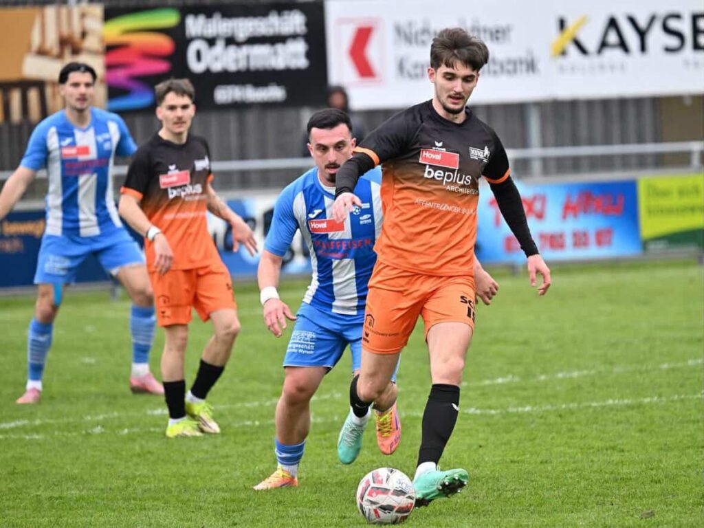 WhatsApp-Image-2026-03-22-at-15.11.391-1024x768 A soccer player in orange and black controls the ball while a blue-and-white defender corners him on a grassy field with advertising boards in the background.