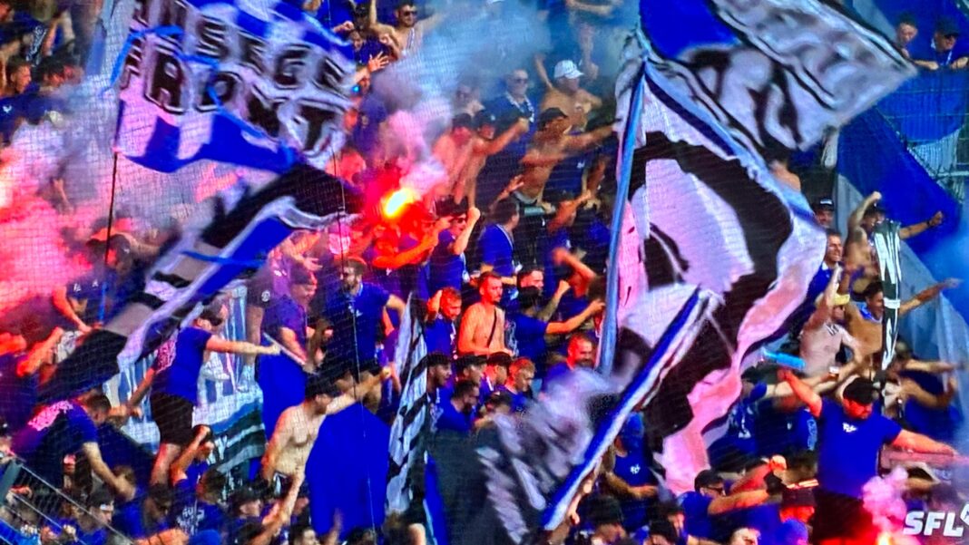 WhatsApp Image 2026-08-09 at 18.47.42 Crowd of blue-clad sports fans cheering with large blue-and-white banners and a flare visible in the stands.