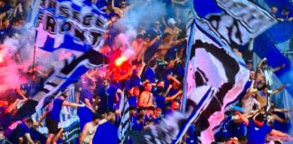 Crowd of blue-clad sports fans cheering with large blue-and-white banners and a flare visible in the stands.