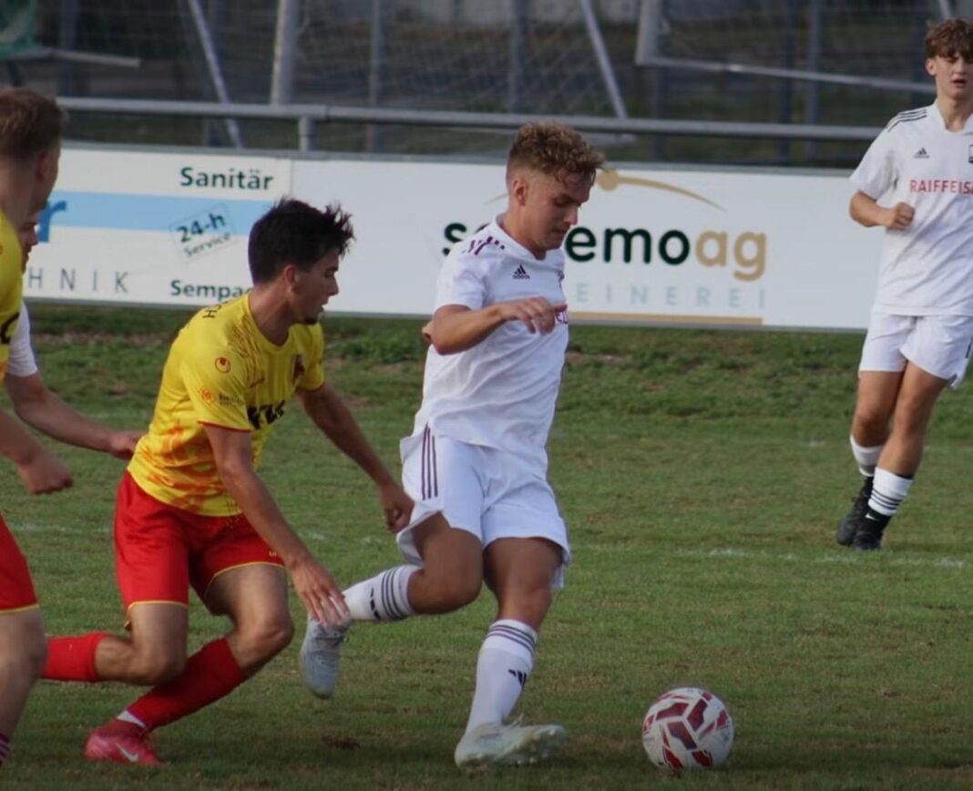 WhatsApp Image 2026-08-24 at 09.45.15 Soccer player in white controls the ball while a yellow-clad opponent closes in on a grassy field with advertising boards in the background.