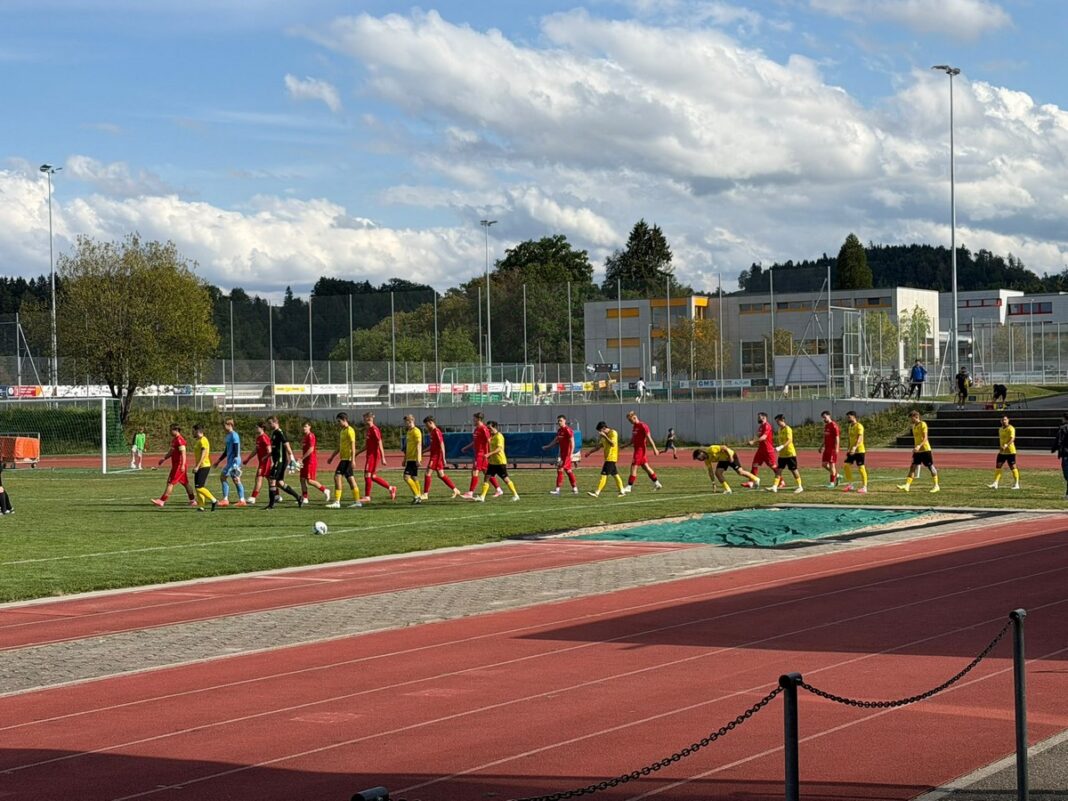WhatsApp Image 2026-08-24 at 14.23.13 (1) Soccer players in red and yellow uniforms walk in a line across a grass field beside a running track.