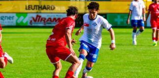 Two soccer players duel for the ball on a green field; one in red kit challenges a white-and-blue kit player as they reach for a dribble.