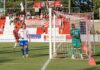 Effiziente Solothurner schlagen Zug 94 Soccer match near the goal: a white-and-blue player stands by the net while a green-clad goalkeeper watches, with cheering fans and banners in the red-and-white stands behind.