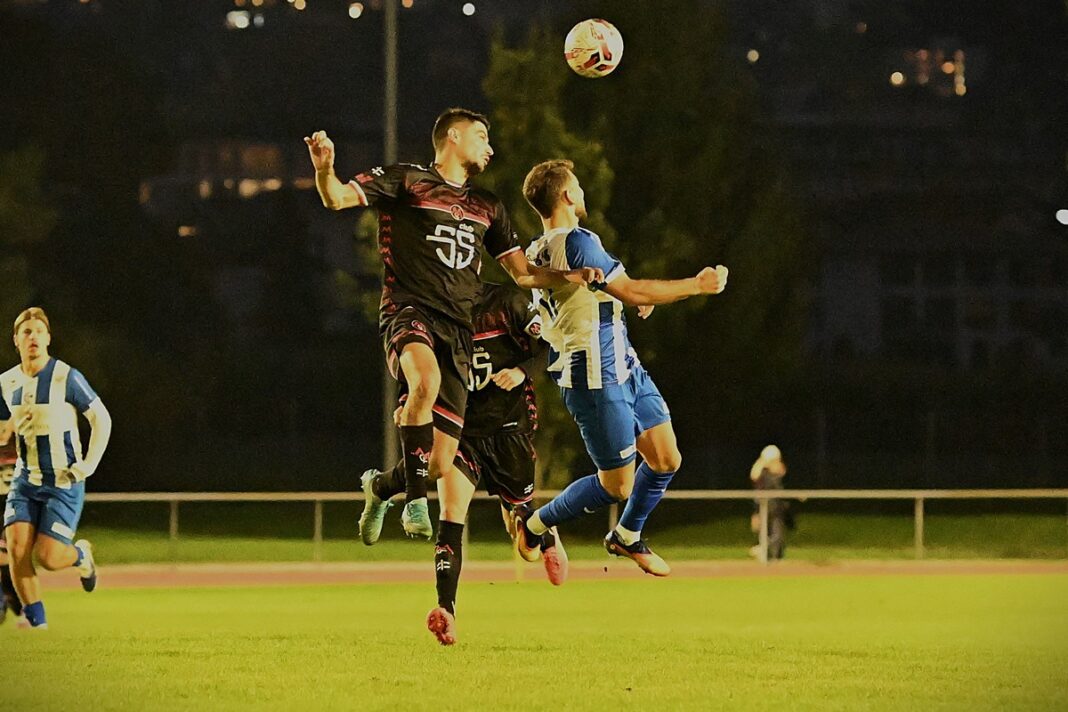 aaaDSC_7935 Two soccer players leap to head the ball during a night match on a grass field, under stadium lights.