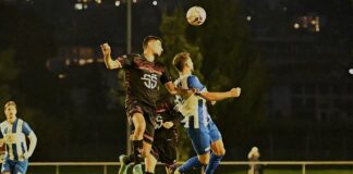 Two soccer players leap to head the ball during a night match on a grass field, under stadium lights.