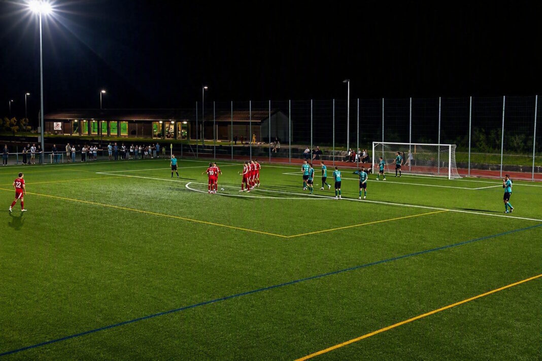 engelbergersc Nighttime soccer match: red team huddles near the center while teal opponents stand nearby under bright floodlights in a fenced stadium area.