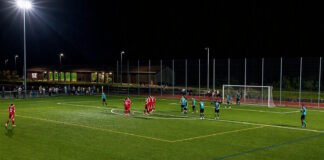 Nighttime soccer match: red team huddles near the center while teal opponents stand nearby under bright floodlights in a fenced stadium area.
