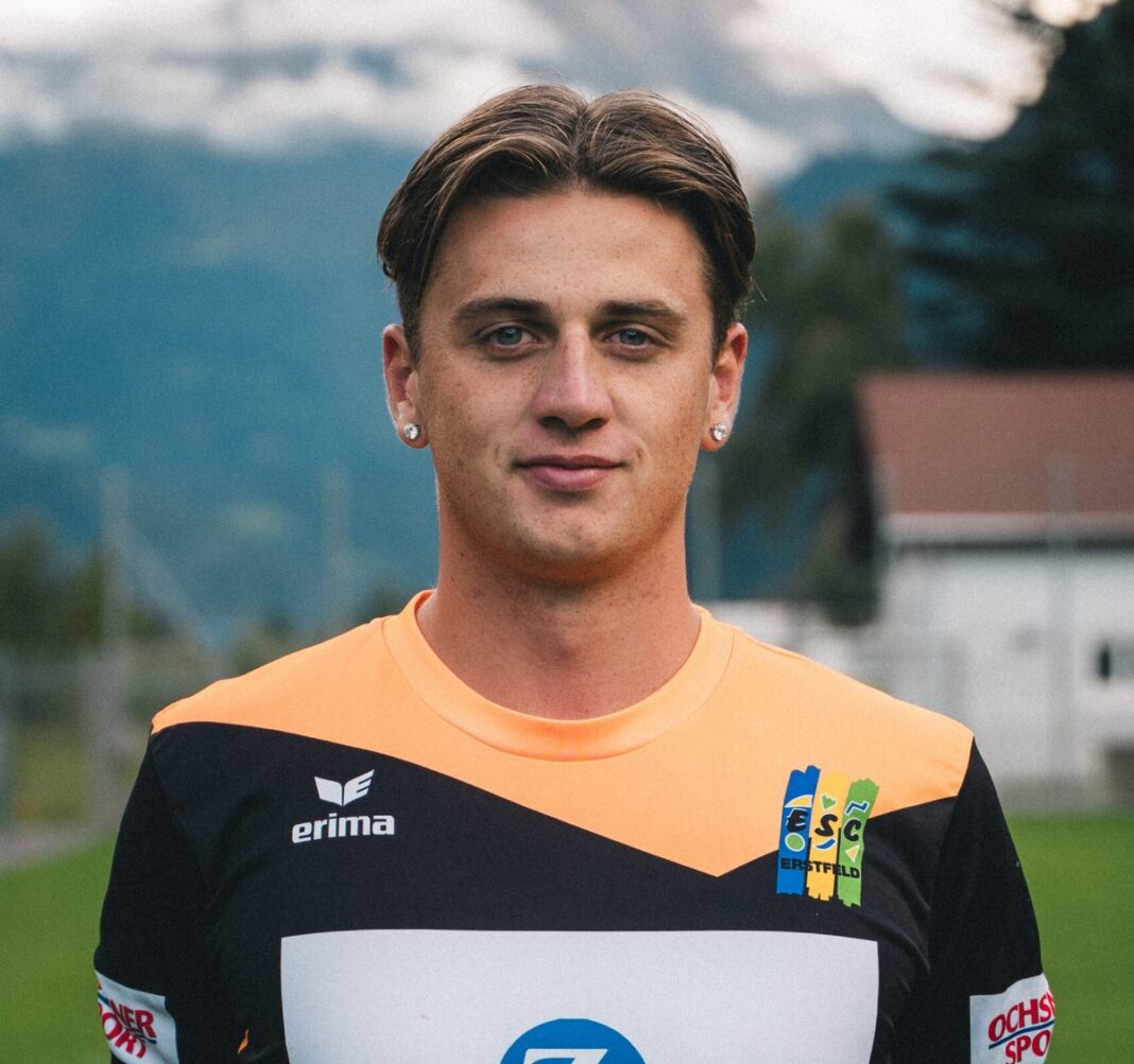 escerstfeld Portrait of a young man in an orange and black sports jersey, standing outdoors with mountains in the background.