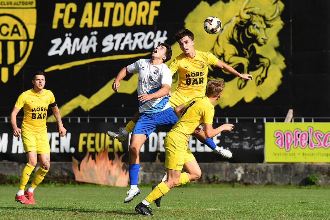fcaltdorf_fcaegeri Two soccer players in yellow jerseys contest the ball midair against a white-and-blue opponent on a sunny grass field, with a large black-and-yellow club banner in the background.
