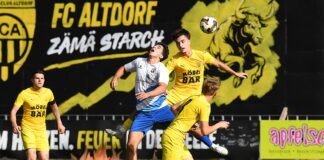 Two soccer players in yellow jerseys contest the ball midair against a white-and-blue opponent on a sunny grass field, with a large black-and-yellow club banner in the background.
