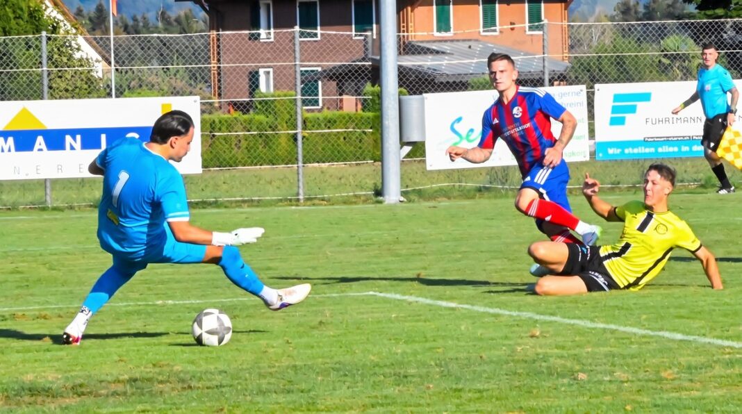 fcbrunnen_fceschenbach Soccer scene: blue-clad goalkeeper about to kick a ball as a yellow-shirted opponent slides in from the right on a green field.