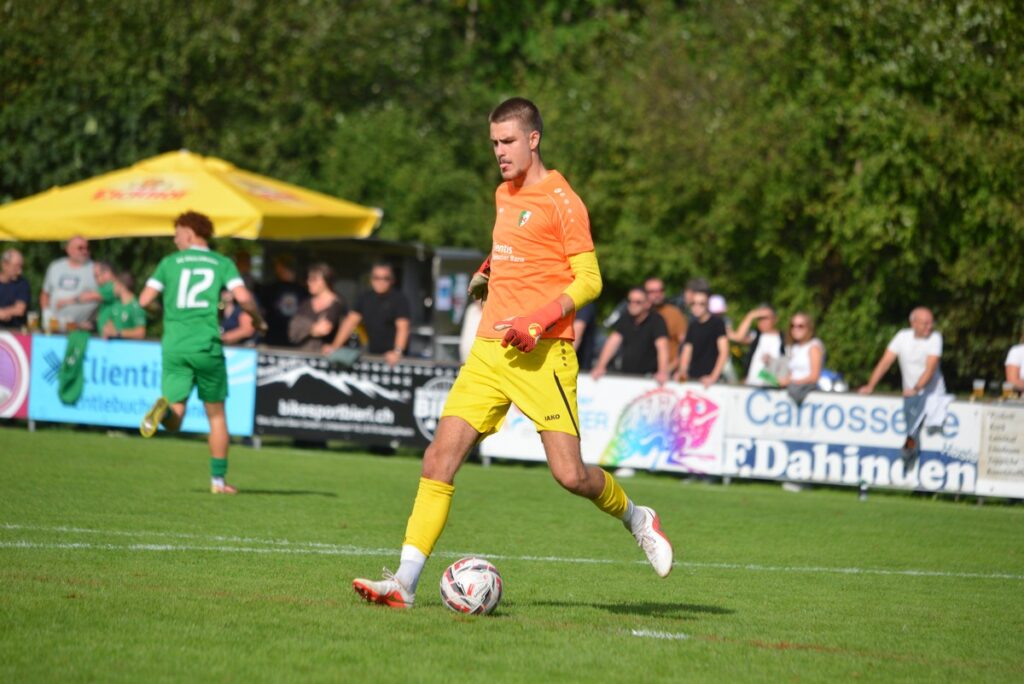 Soccer goalkeeper in orange jersey and yellow shorts dribbling a ball on a sunny green field with spectators and banners behind him.
