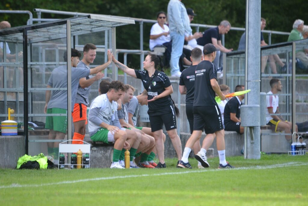 Soccer players on the bench celebrate as a woman in black high-fives teammates in gray shirts.