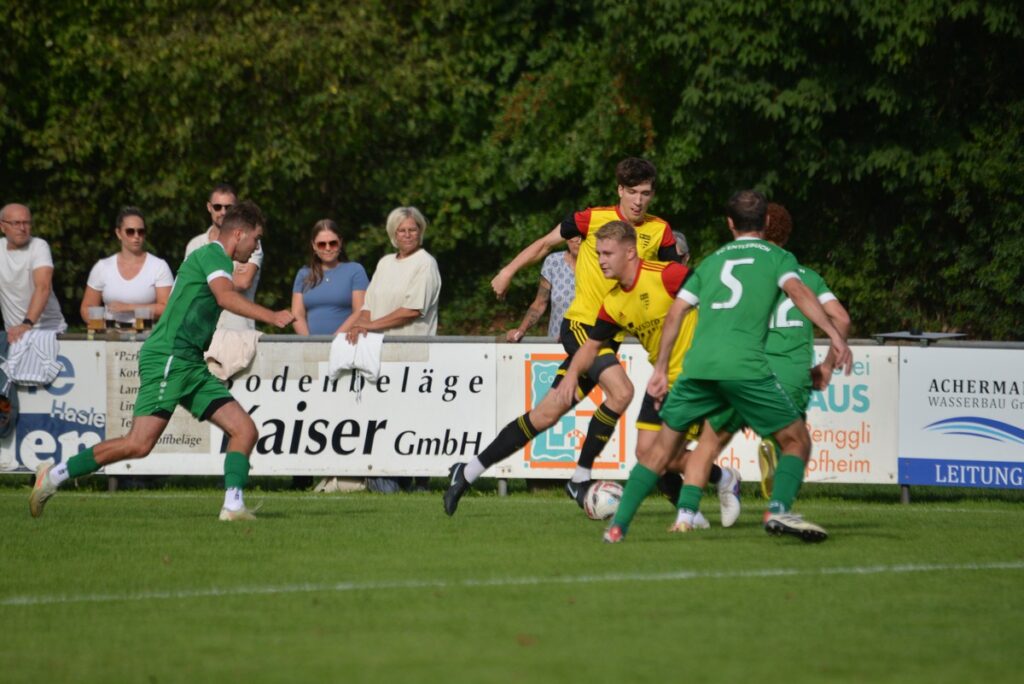 Soccer match action: green and yellow players collide for the ball near the sideline with spectators watching behind advertising boards.