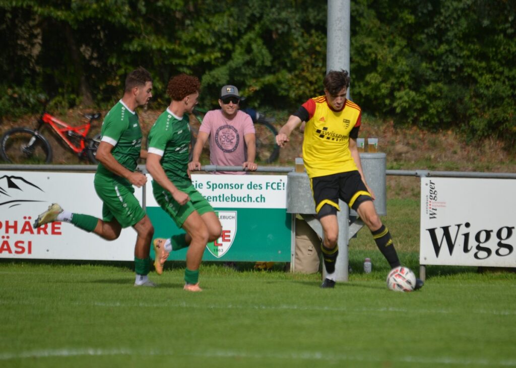Soccer players in green jerseys challenge a player in a yellow jersey near the sideline, ball at his feet on a grassy field with sponsor boards in the background.