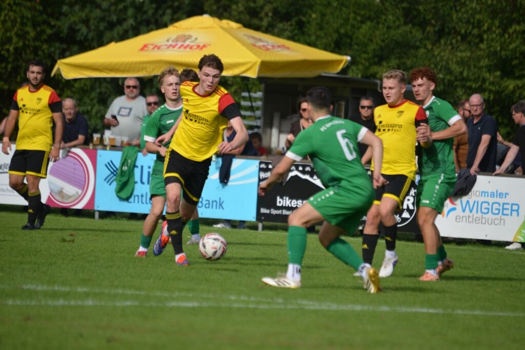 Soccer match with players in yellow and green chasing a ball on a grassy field; spectators and a yellow umbrella in the background.