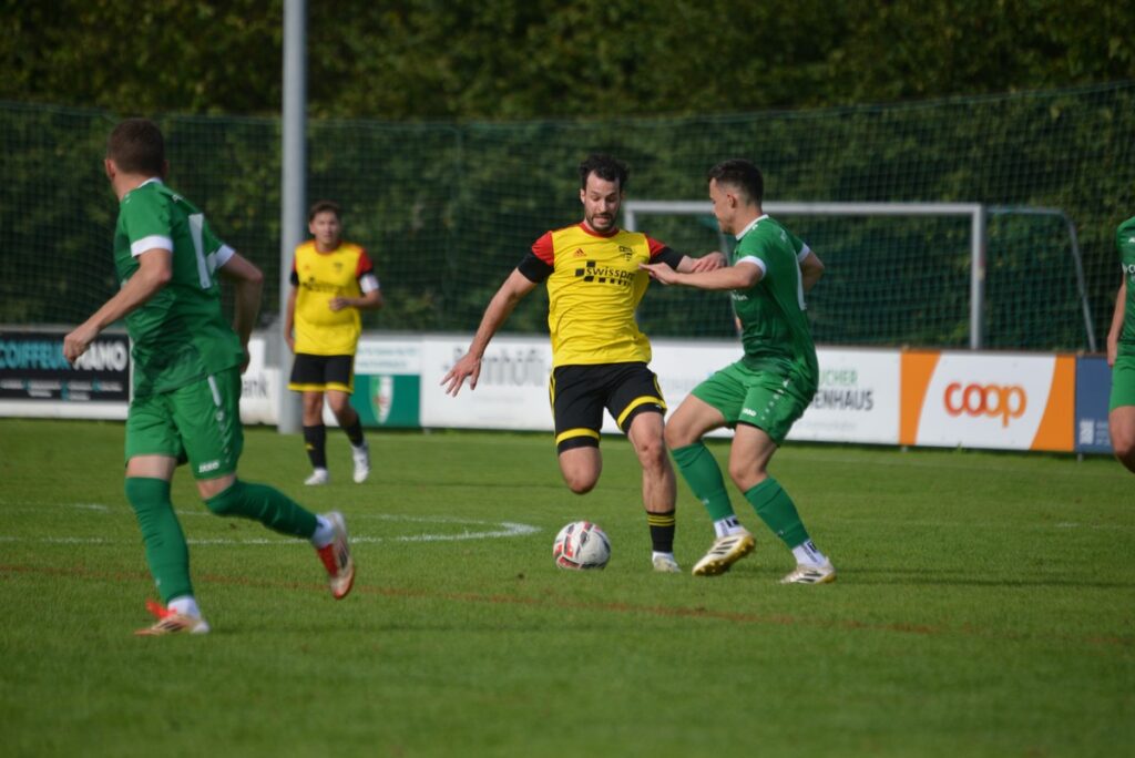 Soccer scene with a yellow-jersey player shielding the ball from a green-jersey opponent on a grass field near advertisements