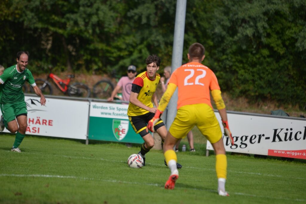 Soccer scene with a yellow-and-black player dribbling the ball toward the goal while a goalkeeper in orange and yellow stands in the foreground.