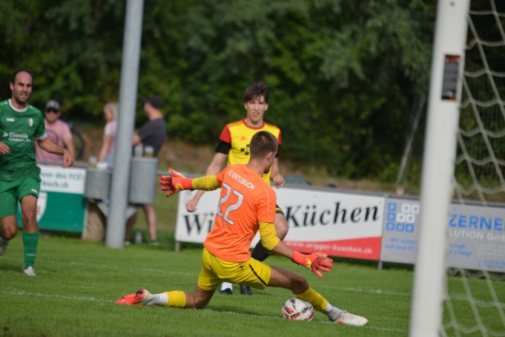 Soccer goalkeeper in orange and yellow dives to block a shot near the goal as a teammate and an opponent watch nearby.