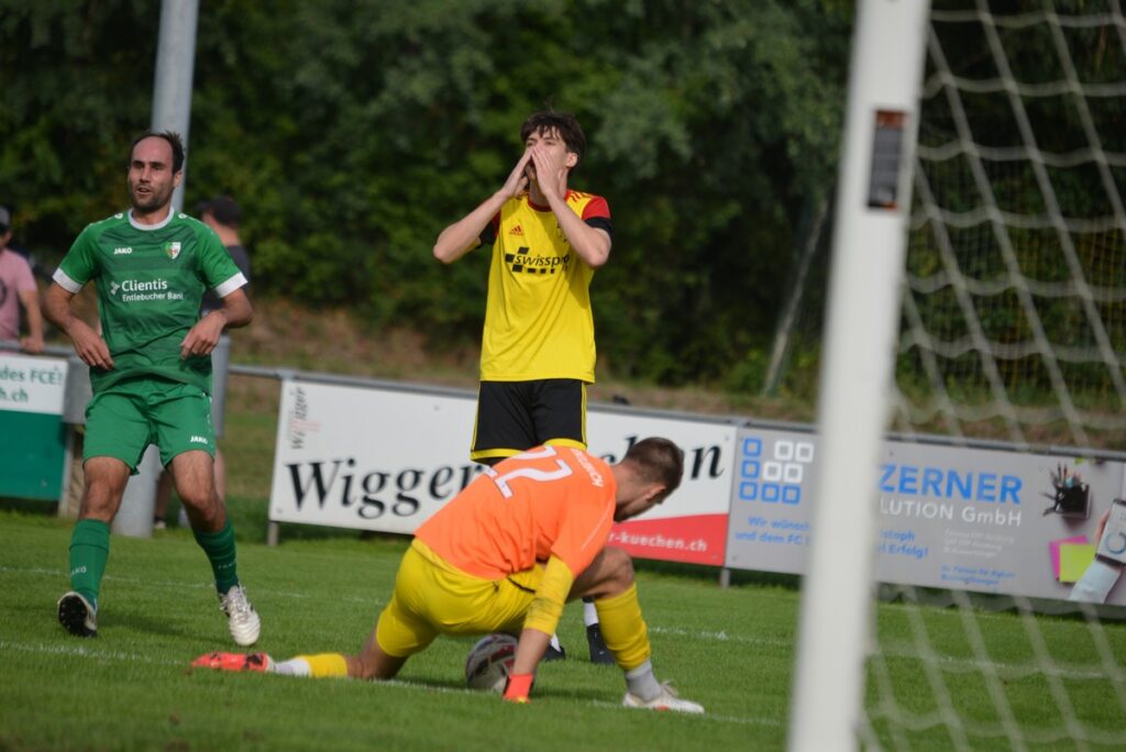 Soccer scene: yellow-jersey player near the goal gesturing, orange-clad goalkeeper on the ground by the ball, and a green-clad defender nearby.