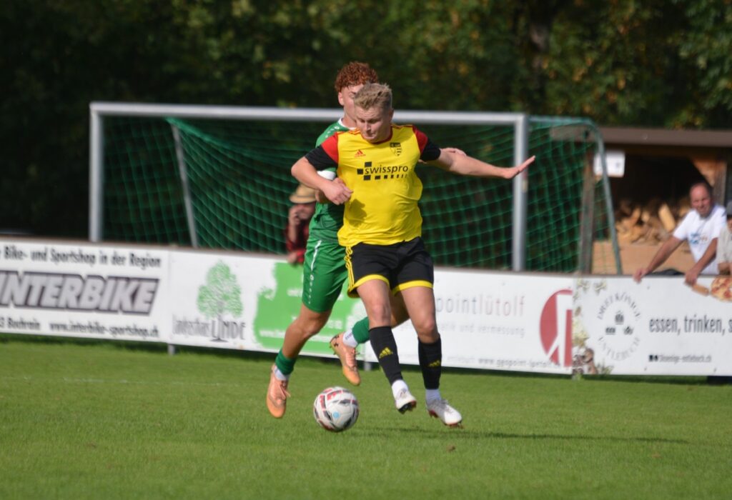 Soccer players contest the ball on a grassy field near the goal, foreground player in yellow and black facing a green defender behind him.