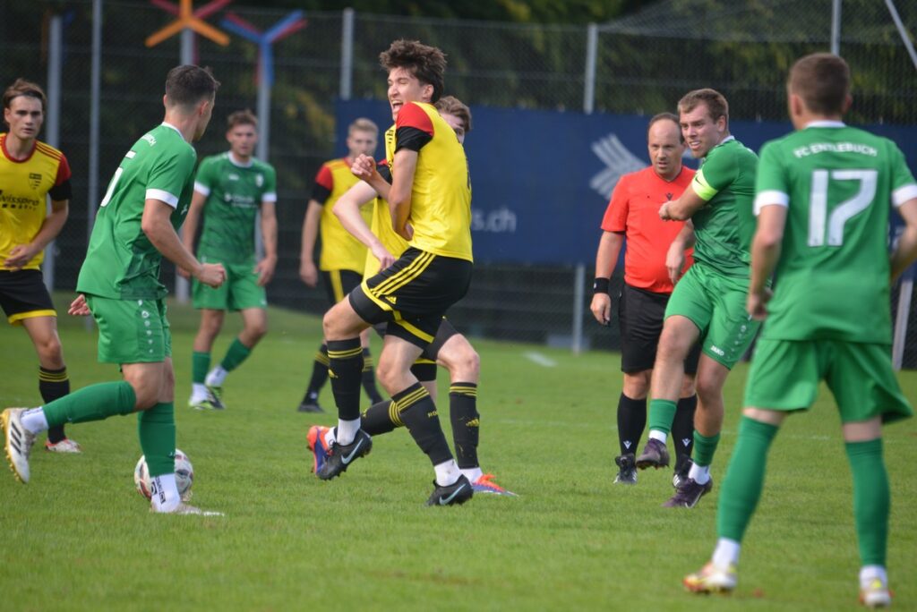 Soccer match with players in green and yellow contesting the ball on a grassy field; a player in yellow leaps while others move around and a referee in orange watches nearby