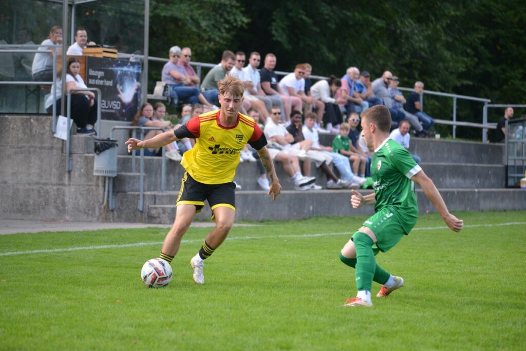 Soccer match: player in yellow jersey dribbles toward goal while a green-clad defender closes in, with spectators watching from the stands behind them on a sunny day.