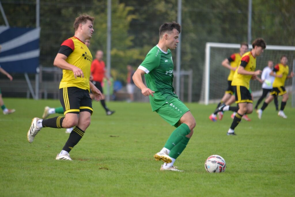 Soccer game: a green-team player dribbles the ball while yellow-team players chase nearby on a grassy field near the goal area.
