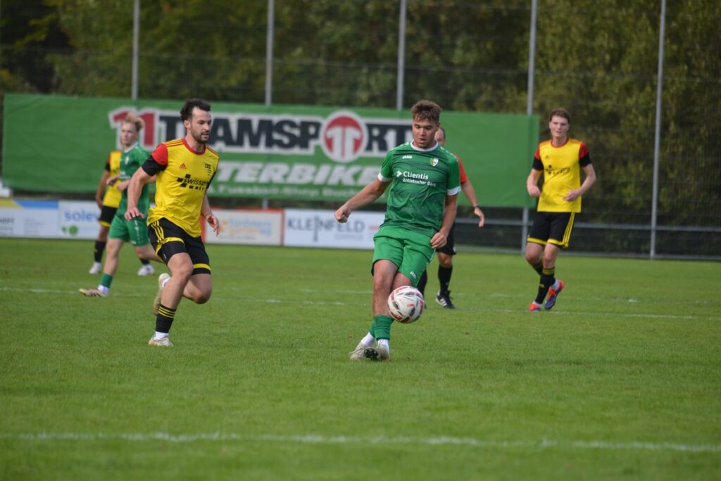 Soccer match: green-clad player controls the ball while a yellow-jersey opponent approaches on a grassy field behind a advertising barrier.