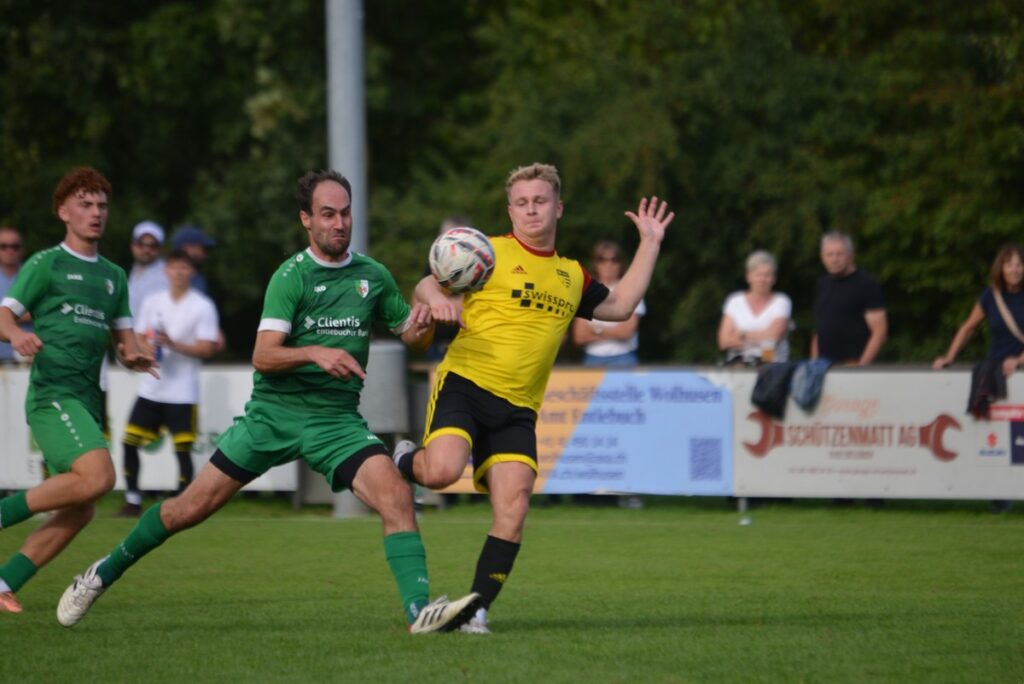 Soccer player in yellow shoots or traps the ball while a green-clad defender reaches in, on a grassy field with spectators in the background.