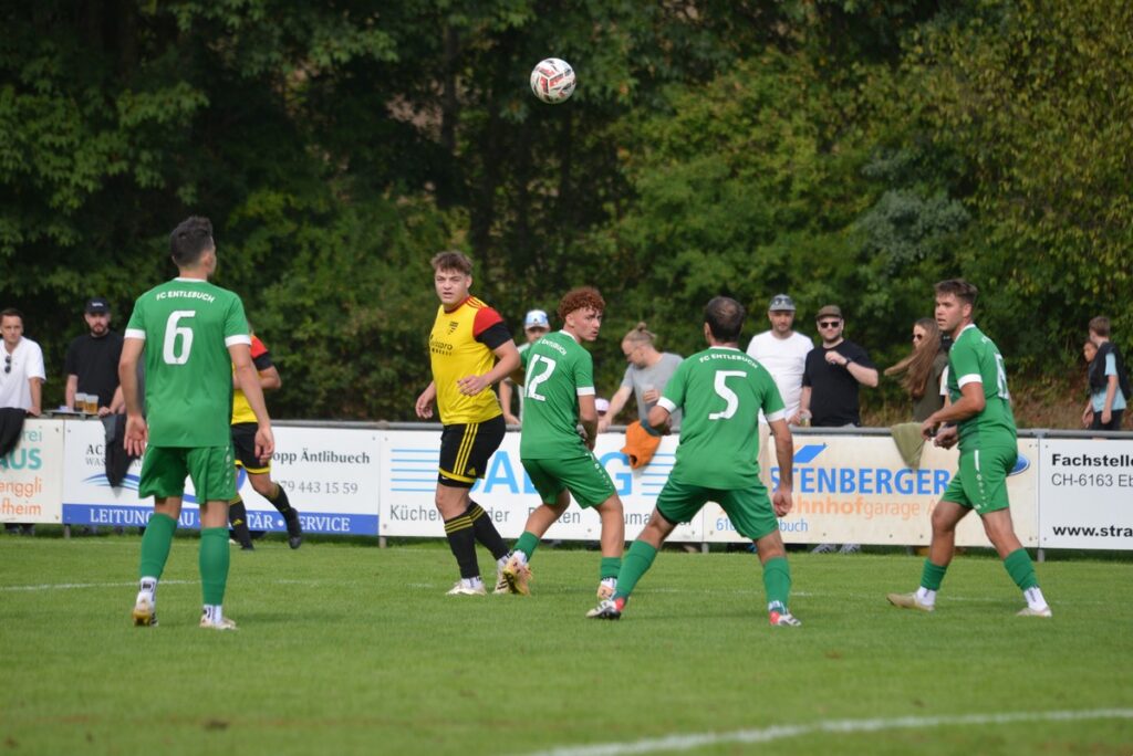 Soccer players chase the ball in midair during a match; green team vs yellow-black team on a grassy field with spectators behind a barrier.
