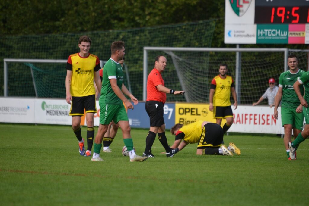 Soccer match on a green field with players in yellow and green uniforms; the referee in orange gestures while a player lies on the ground nearby.
