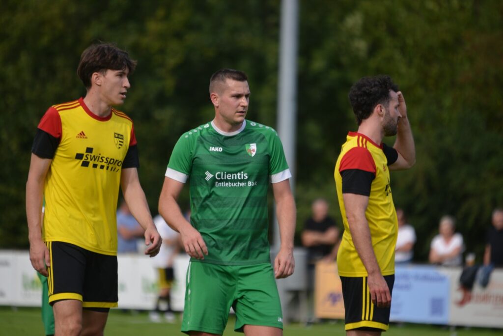 Three soccer players on a field: one in green center, two in yellow jerseys on the sides, likely mid-game action.