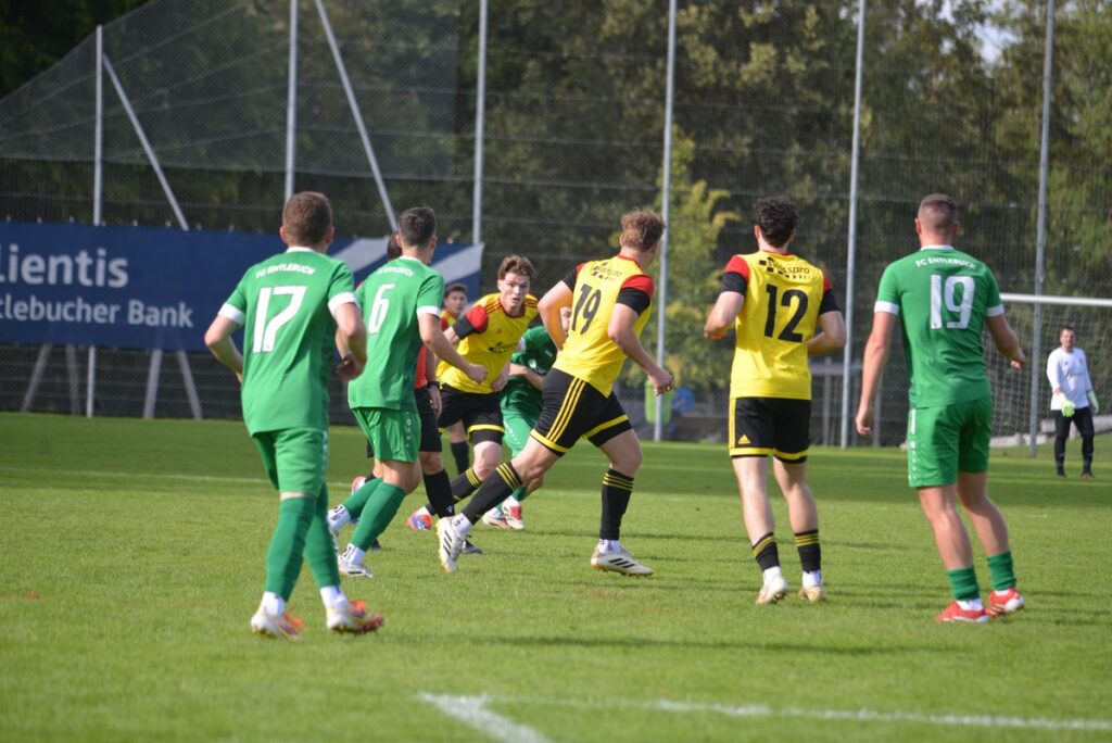 Soccer players in green and yellow jerseys chase the ball on a sunny grassy field, with a goalie near the goal in the background.