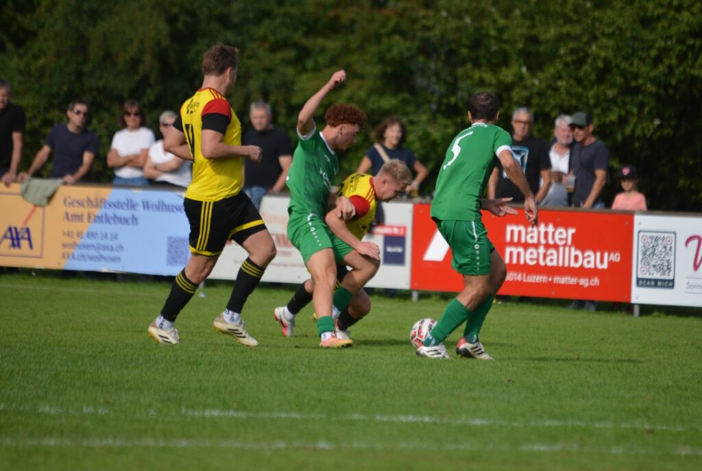 Soccer players in green and yellow jerseys contest the ball on a sunny field, with spectators along the sideline and advertising boards behind them.