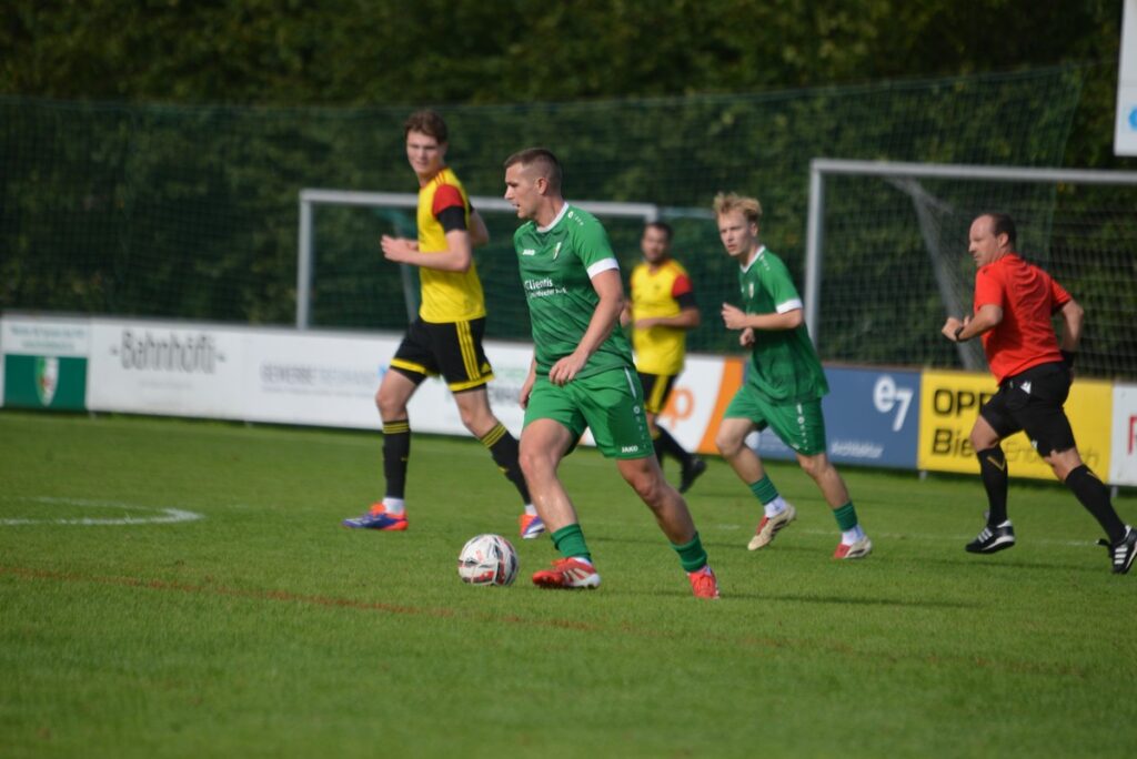 Soccer player in green controls the ball while teammates and opponents run across a grassy pitch during a daytime match.