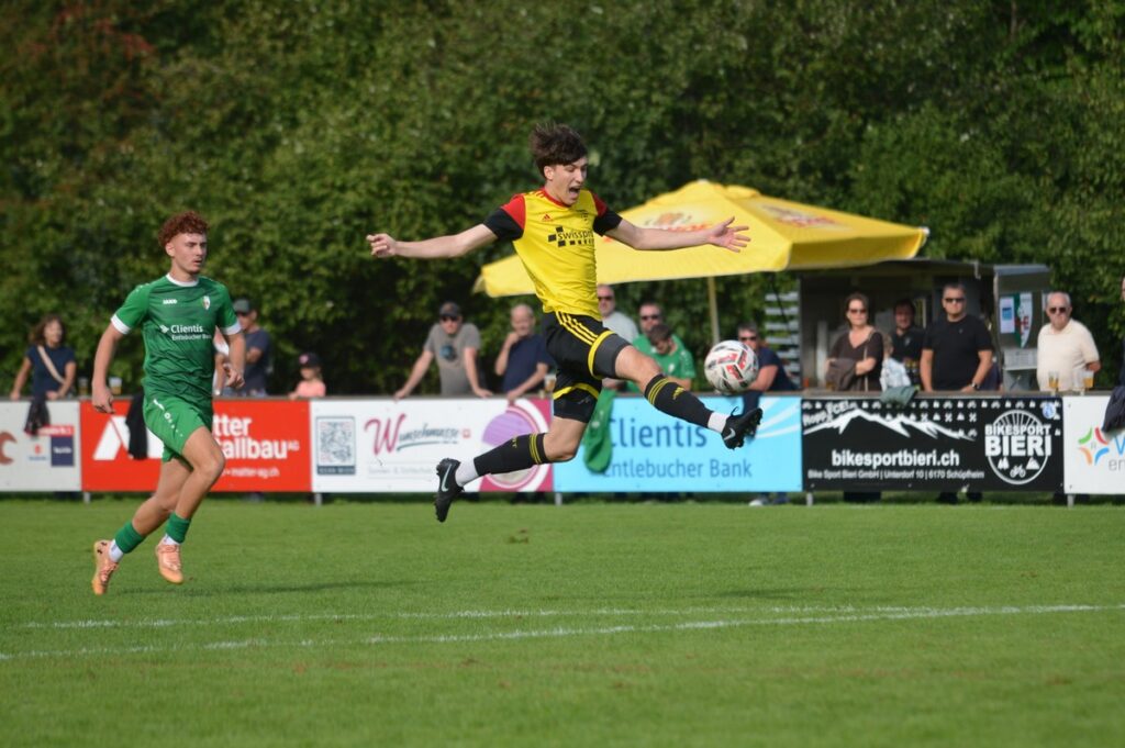 Soccer scene: yellow-shirted player leaps midair to kick a ball as a green-clad opponent watches on a grassy field with spectators and banners in the background.