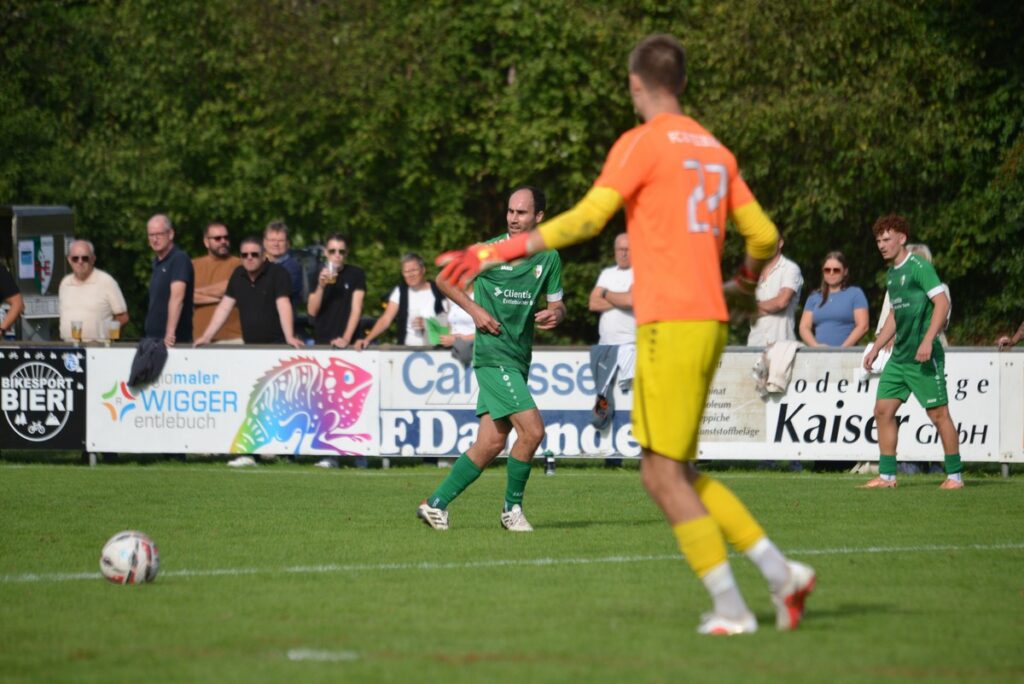 Soccer scene: a green team player with ball near the sideline while an orange-clad goalkeeper stands in foreground and spectators watch behind advertising boards.