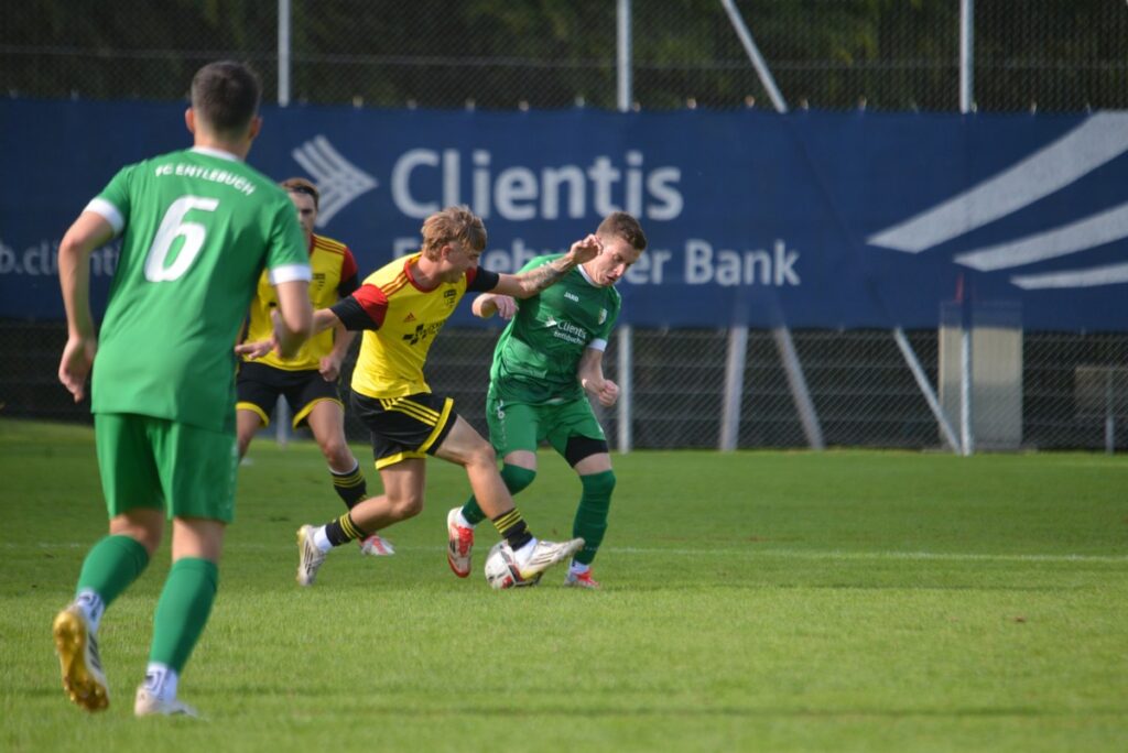 Two soccer players in yellow and green contest a ball on a grass field during a match, with a blue banner in the background.